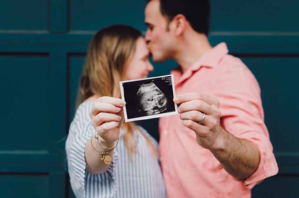 An expecting couple hold the photo of their unborn babies sonogram while the man kisses the woman on the forehead in the background. trying to conceive