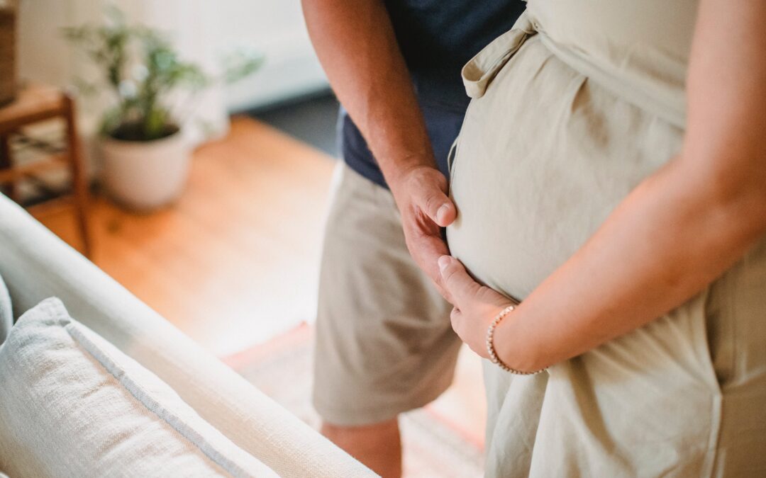 pregnant woman holding her belly while her partner places a hand on her stomach as well. fertility coaching, fertility support, infertility